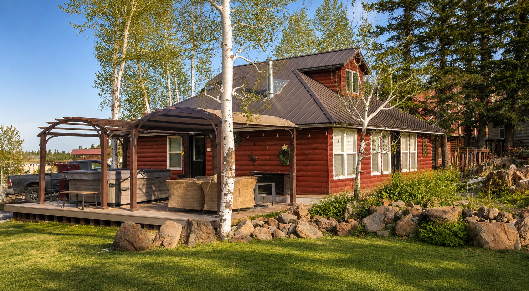 A Lazy Duck log cottage with its deck, pergola, and hot tub framed by aspens and a green mountain lawn